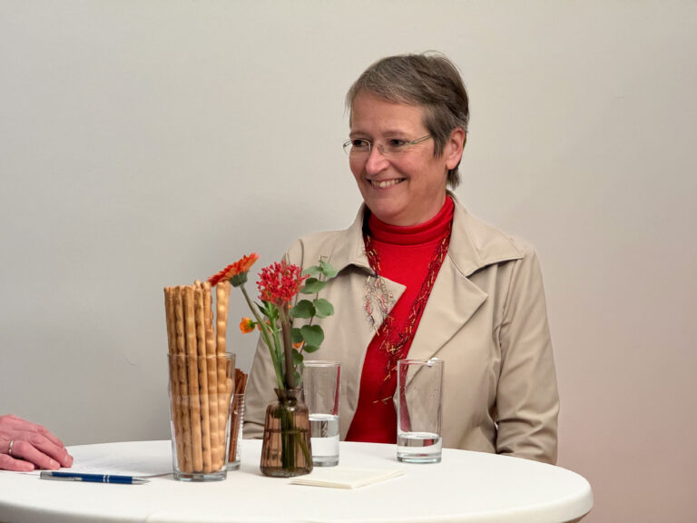 Monika Auer steht während ihres Dialoges mit Hemma Bieser hinter einem Tisch. Auf dem Tisch stehen Grissini und Blumen.